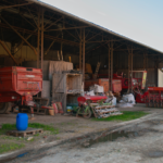 Vue d'un hangar attenant à une ferme, où attendent de nombreux outils et engins agricoles, stockés à côtés de bottes de foin.