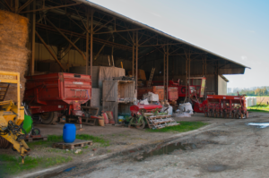 Vue d'un hangar attenant à une ferme, où attendent de nombreux outils et engins agricoles, stockés à côtés de bottes de foin.