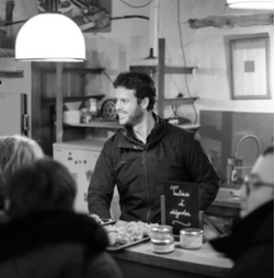 Portrait photo de Mathieu Bordelet traiteur, lors d'un événement. Mathieu se tient derrière le comptoir sur lequel il a disposé ses créations. Des personnes attendent pour se servir.