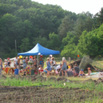 Une quarantaine de personnes sont réunies pour une fête à côté de champs cultivés. Des tables sont dressées et un petit chapiteau a été monté pour les abriter de la pluie ou du soleil. Au fond, une forêt.