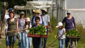 Un groupe d'adultes et d'enfants portent des outils et des plants afin de les planter lors d'un chantier participatif organisé par une AMAP.