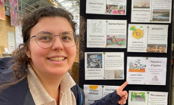 Portrait de l'entrepreneuse Agathe Villien, productrice de plants. Elle pose devant un présentoir qui indique sa pépinière lors d'un événement.