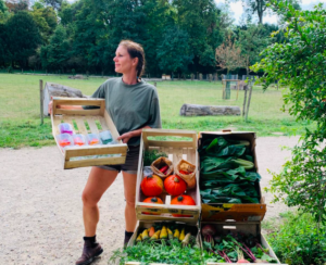 Une maraîchère tient une cagette dans les mains et pose devant un chariot rempli de cagettes de récolte de légumes variés