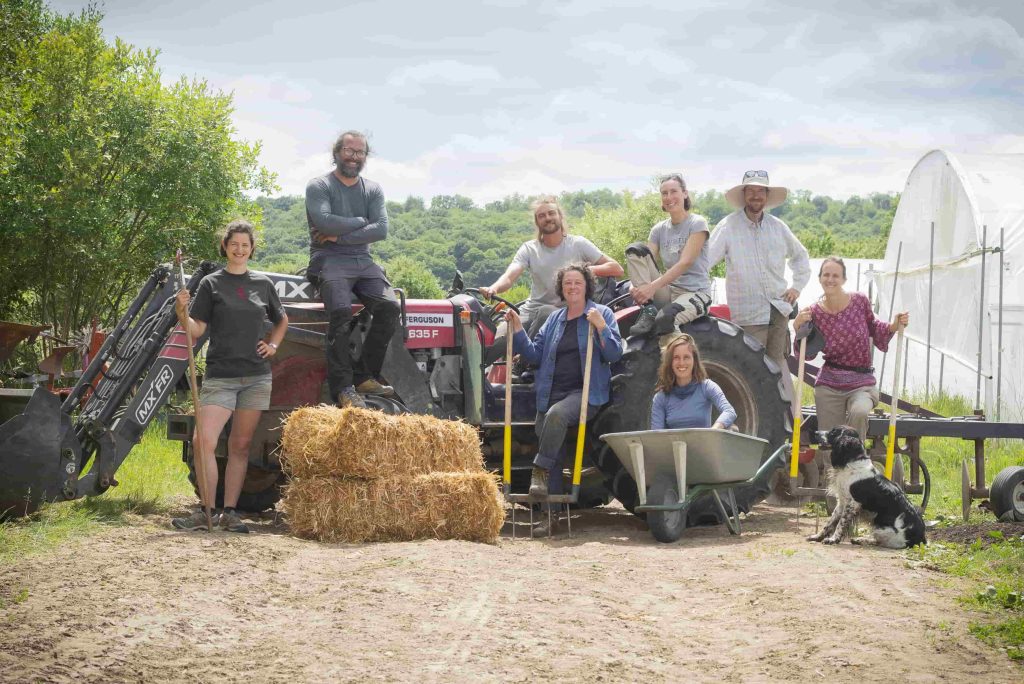 Une dizaine de personnes posent avec leurs outils et des bottes de foin à côté de serres maraîchères